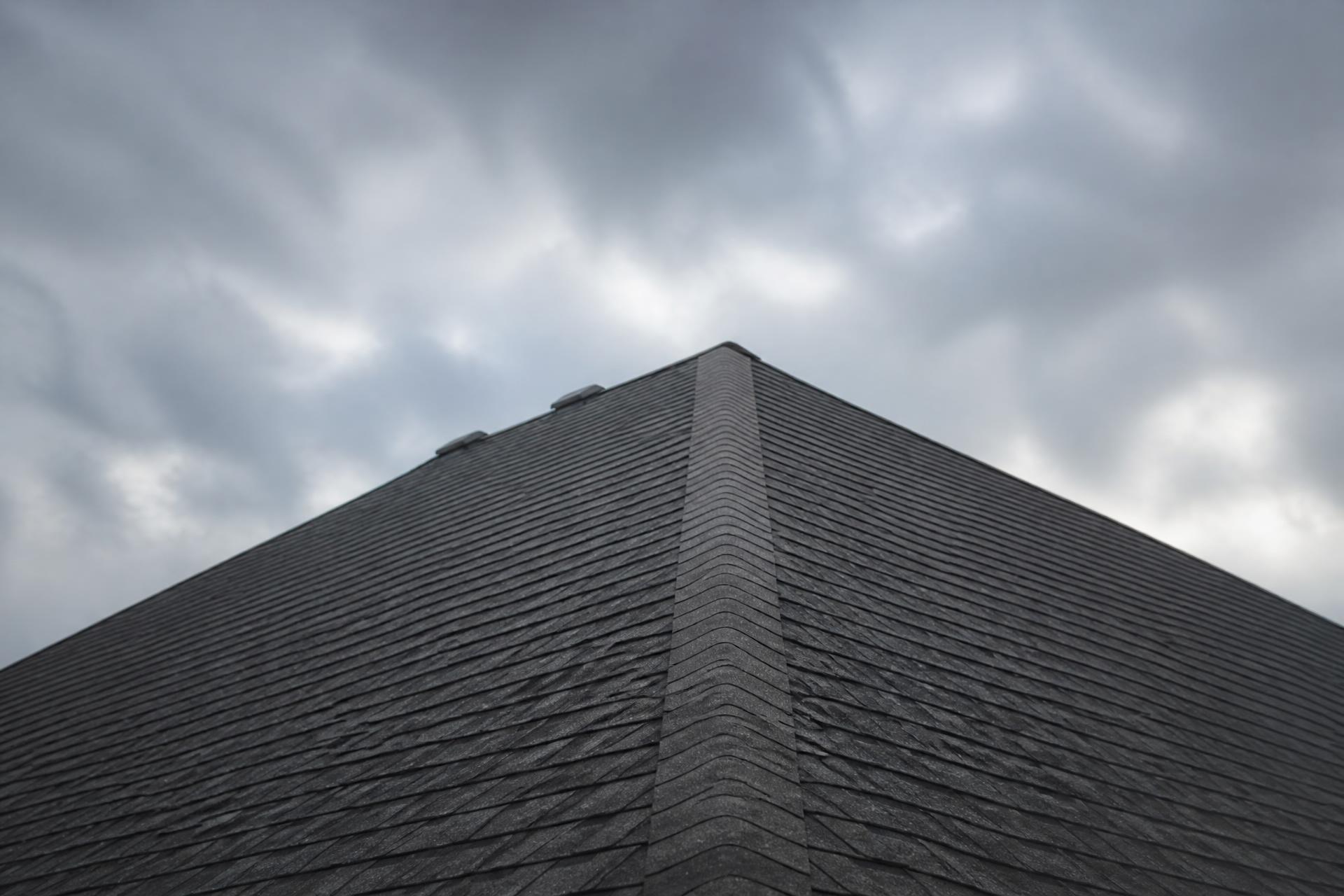 Dark grey shingled hip roof with ridge lines converging against a cloudy sky